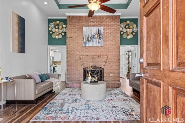 living area featuring ceiling fan, dark wood-style flooring, a brick fireplace, and crown molding