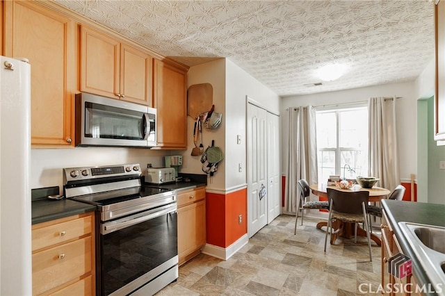 kitchen with baseboards, dark countertops, appliances with stainless steel finishes, a textured ceiling, and light brown cabinetry