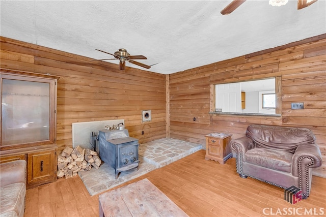 living room with ceiling fan, a textured ceiling, wood walls, wood finished floors, and a wood stove