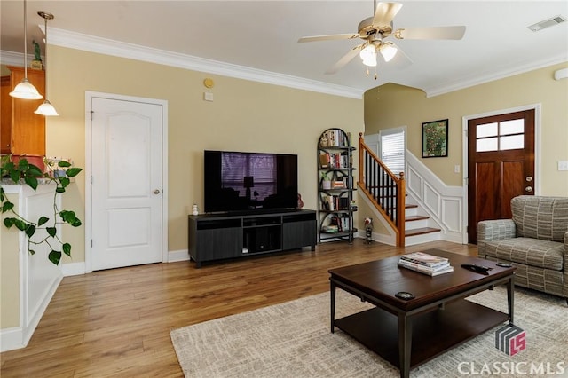 living room with light wood-style flooring, a ceiling fan, visible vents, stairway, and crown molding