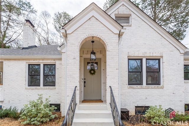 view of front of home with a shingled roof, crawl space, brick siding, and a chimney