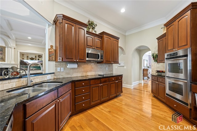 kitchen with light wood finished floors, arched walkways, dark stone counters, stainless steel appliances, and a sink