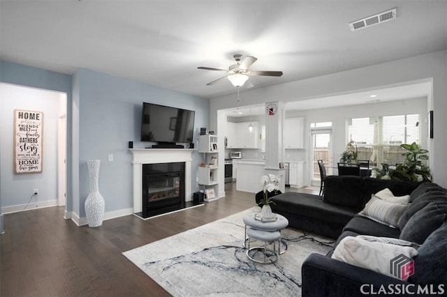 living area featuring visible vents, a glass covered fireplace, dark wood finished floors, baseboards, and ceiling fan