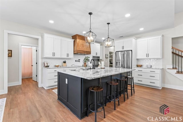 kitchen with light wood finished floors, custom exhaust hood, stainless steel fridge with ice dispenser, a kitchen island with sink, and white cabinets