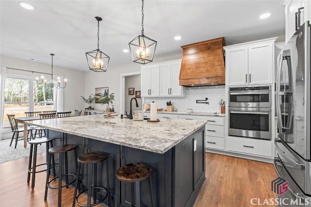 kitchen featuring wood finished floors, custom exhaust hood, white cabinets, appliances with stainless steel finishes, and backsplash