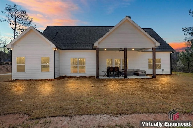 back of house at dusk featuring a lawn, a ceiling fan, roof with shingles, an outdoor hangout area, and a patio area