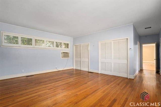 unfurnished bedroom with visible vents, baseboards, a wall mounted AC, hardwood / wood-style flooring, and multiple closets