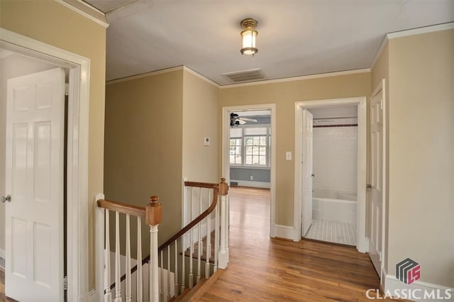 hallway with wood finished floors, visible vents, baseboards, crown molding, and an upstairs landing