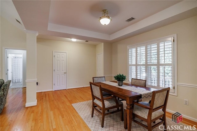 dining space featuring visible vents, light wood-style flooring, baseboards, a raised ceiling, and ornate columns