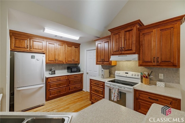 kitchen featuring under cabinet range hood, white appliances, brown cabinetry, light countertops, and lofted ceiling