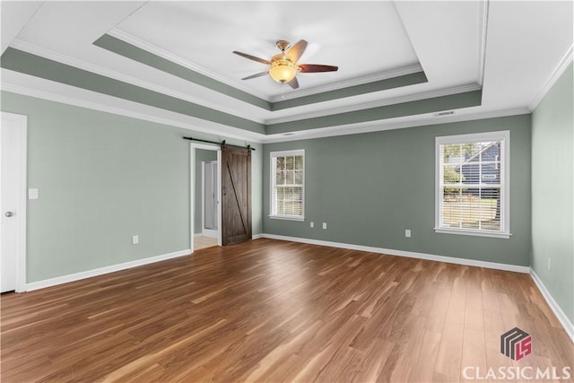 empty room with a ceiling fan, wood finished floors, baseboards, a tray ceiling, and a barn door