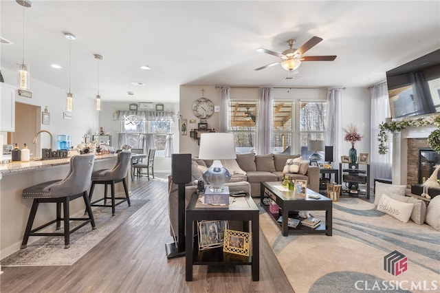 living area featuring light wood finished floors, a glass covered fireplace, a healthy amount of sunlight, and a ceiling fan