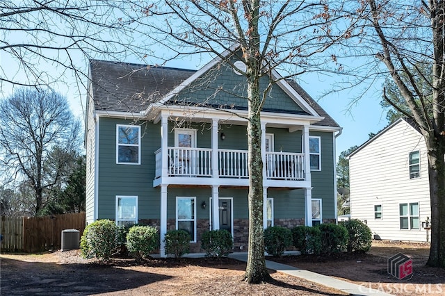 view of front facade with fence, roof with shingles, cooling unit, a balcony, and stone siding