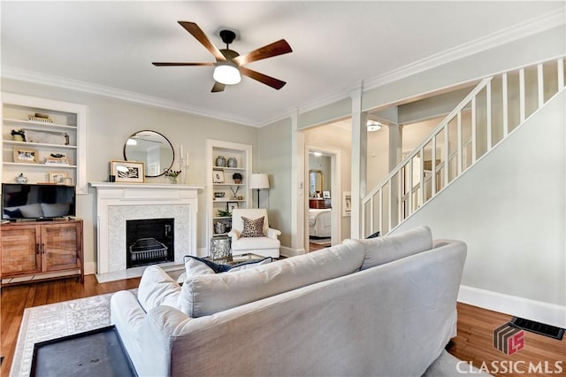 living area with ceiling fan, a fireplace with flush hearth, built in features, wood finished floors, and ornamental molding