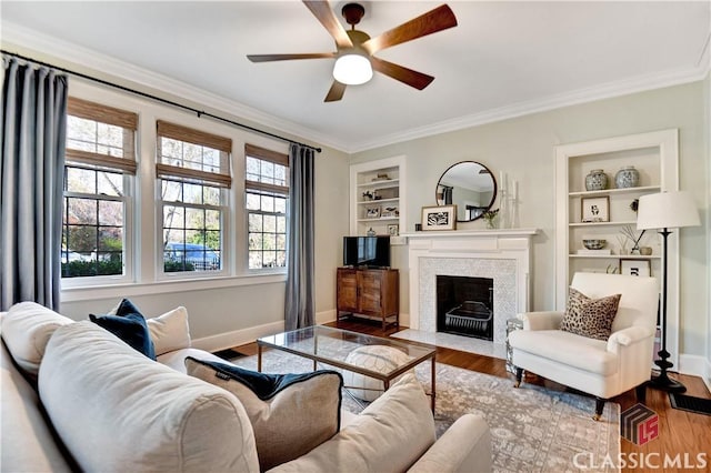living room featuring built in shelves, a ceiling fan, baseboards, wood finished floors, and ornamental molding