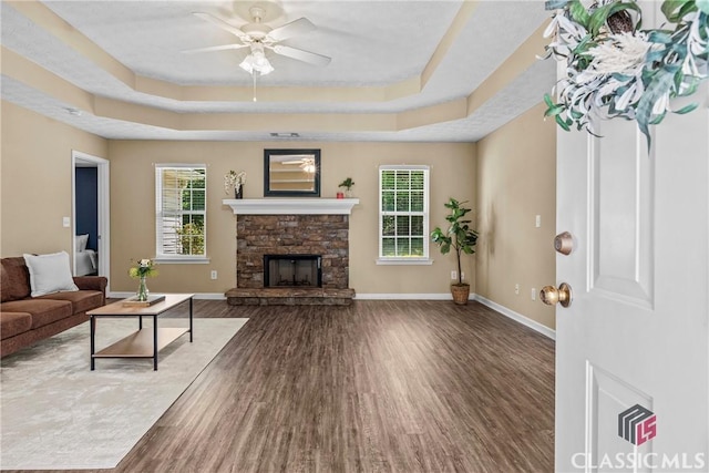 living room with a raised ceiling, wood finished floors, a ceiling fan, baseboards, and a stone fireplace