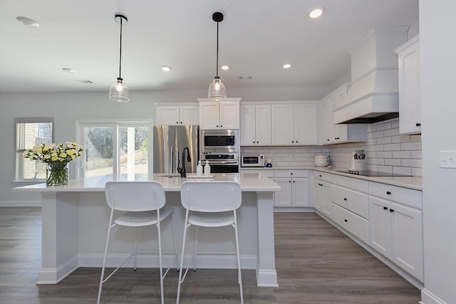 kitchen with premium range hood, a kitchen island with sink, backsplash, stainless steel appliances, and light countertops