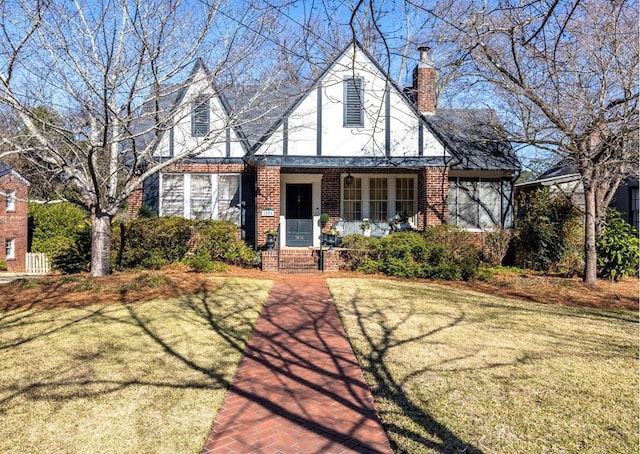 tudor house with a chimney, an attached garage, covered porch, a front lawn, and brick siding