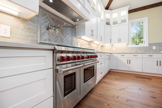 kitchen with custom exhaust hood, backsplash, white cabinets, range with two ovens, and light hardwood / wood-style floors