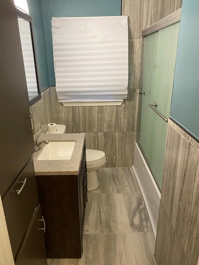 full bathroom with shower / bath combination with glass door, vanity, toilet, and tile walls