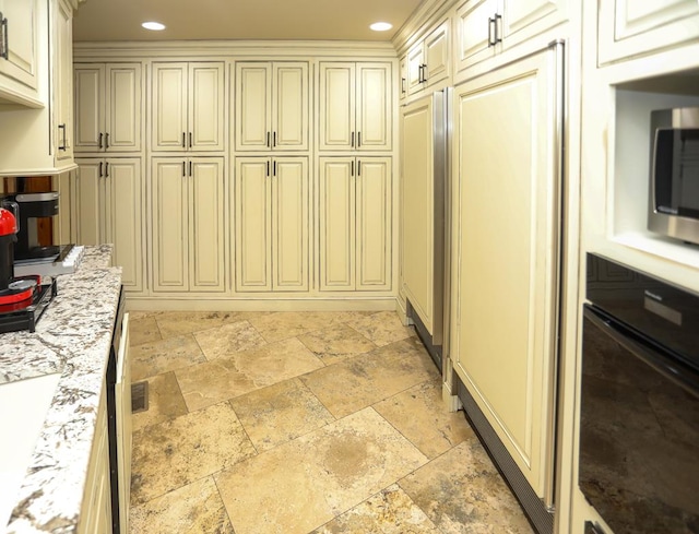 kitchen with cream cabinetry, oven, light stone counters, and crown molding