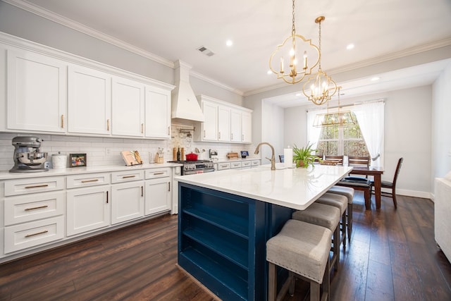 kitchen with a kitchen island with sink, hanging light fixtures, custom range hood, and white cabinets