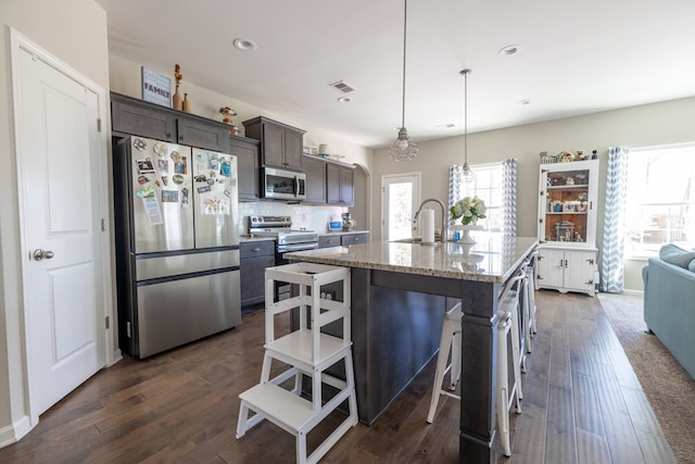 kitchen featuring light stone counters, stainless steel appliances, dark wood-type flooring, dark brown cabinetry, and an island with sink