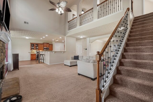 living room with stairway, visible vents, ceiling fan, a stone fireplace, and light carpet