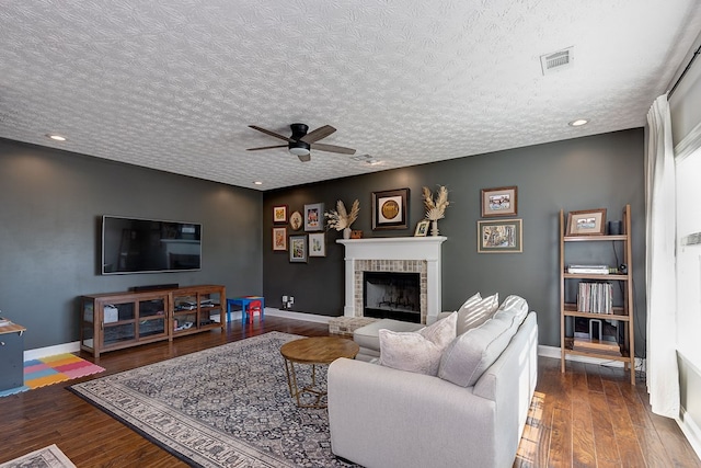 living room with dark wood-type flooring, a brick fireplace, a textured ceiling, and ceiling fan