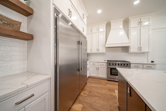 kitchen with light stone counters, white cabinets, high quality appliances, light wood-type flooring, and premium range hood