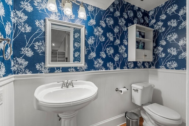 bathroom with a sink, a wainscoted wall, toilet, and wallpapered walls