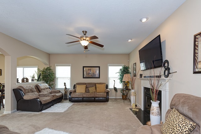 carpeted living room featuring arched walkways, ceiling fan, a fireplace with flush hearth, and baseboards