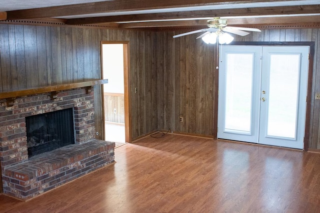 unfurnished living room featuring beam ceiling, ceiling fan, a brick fireplace, wood-type flooring, and wooden walls