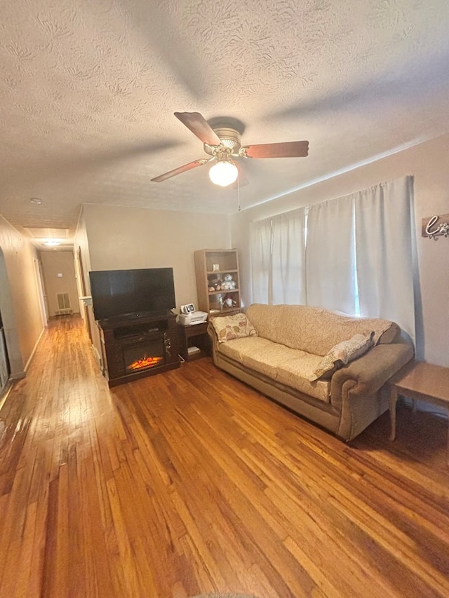 living room featuring a ceiling fan, a warm lit fireplace, a textured ceiling, and hardwood / wood-style floors