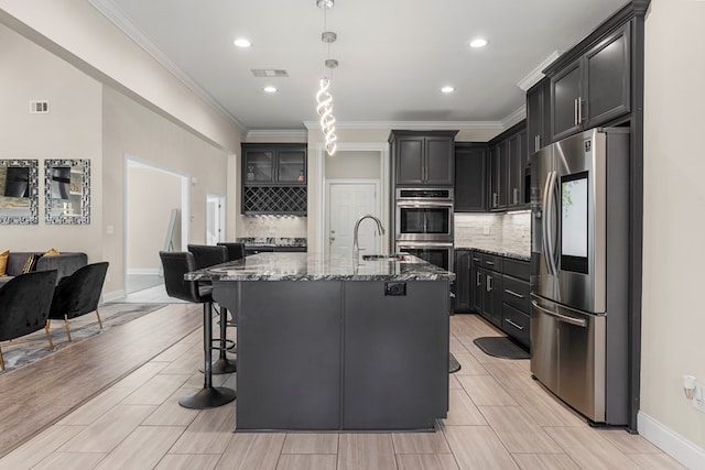 kitchen featuring decorative backsplash, ornamental molding, stainless steel appliances, a kitchen island with sink, and stone counters