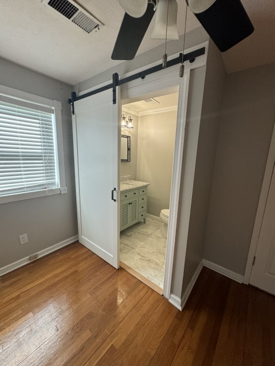 unfurnished bedroom with ceiling fan, a barn door, ensuite bathroom, wood-type flooring, and ornamental molding