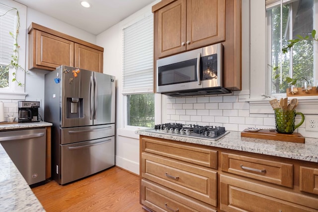 kitchen with brown cabinetry, decorative backsplash, light wood-style floors, appliances with stainless steel finishes, and a wealth of natural light