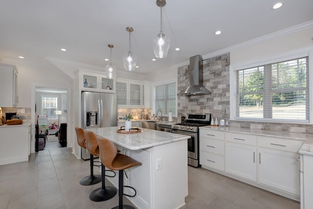 kitchen with wall chimney range hood, a wealth of natural light, appliances with stainless steel finishes, a kitchen island, and white cabinetry