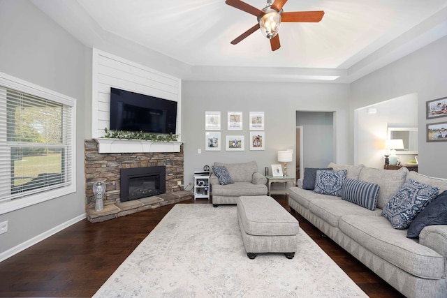 living area with a ceiling fan, dark wood-style floors, a fireplace, a raised ceiling, and baseboards
