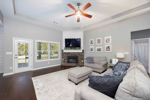 living room featuring ceiling fan, dark wood finished floors, a tray ceiling, a stone fireplace, and baseboards