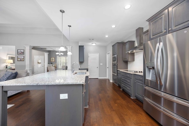 kitchen featuring appliances with stainless steel finishes, open floor plan, a sink, wall chimney range hood, and light stone counters