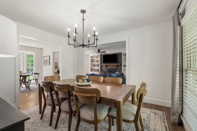 dining room featuring baseboards, wood finished floors, ceiling fan with notable chandelier, and a brick fireplace