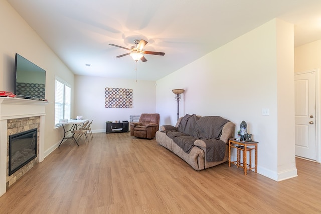 living room featuring light wood-style floors, ceiling fan, a stone fireplace, and baseboards