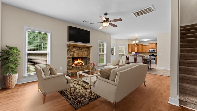 living room featuring ceiling fan with notable chandelier, light wood-type flooring, a stone fireplace, and a healthy amount of sunlight