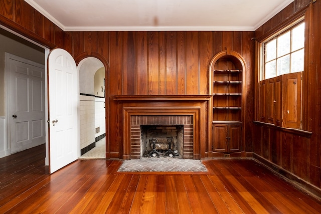 unfurnished living room with crown molding, a brick fireplace, wooden walls, hardwood / wood-style flooring, and arched walkways