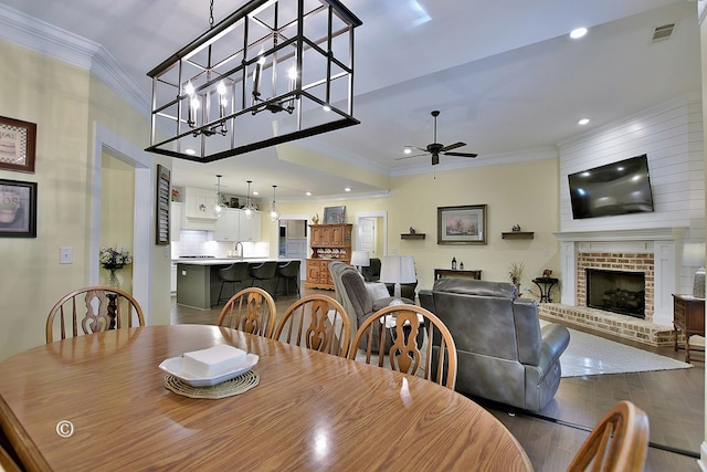 dining space featuring wood-type flooring, ceiling fan with notable chandelier, a brick fireplace, and ornamental molding