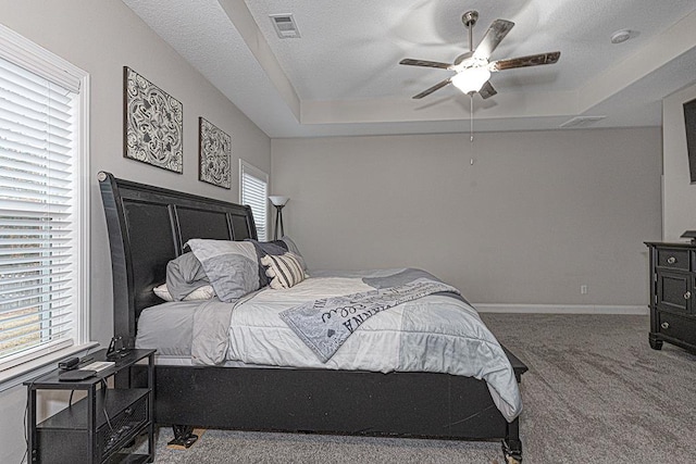 carpeted bedroom with a textured ceiling, ceiling fan, multiple windows, and a tray ceiling