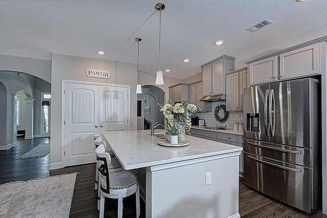 kitchen with pendant lighting, backsplash, dark wood-type flooring, stainless steel refrigerator with ice dispenser, and an island with sink