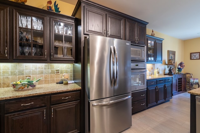 kitchen featuring light stone counters, dark brown cabinetry, stainless steel appliances, decorative backsplash, and glass insert cabinets