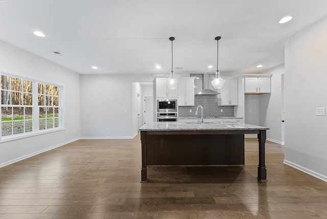 kitchen with light stone countertops, white cabinetry, wall chimney exhaust hood, stainless steel appliances, and a kitchen island with sink